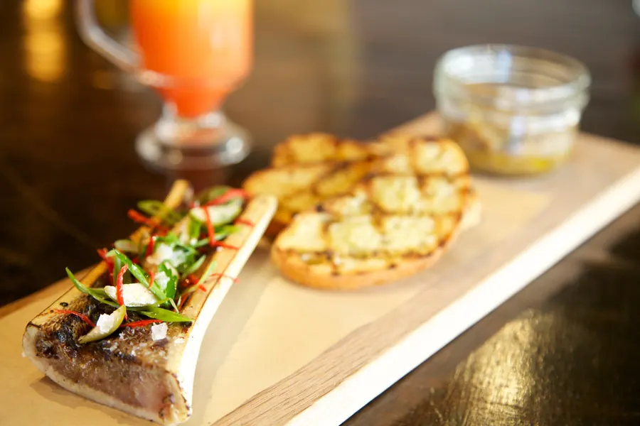 Roasted bone marrow with grilled toast on a wooden board, a small jar and an orange drink behind