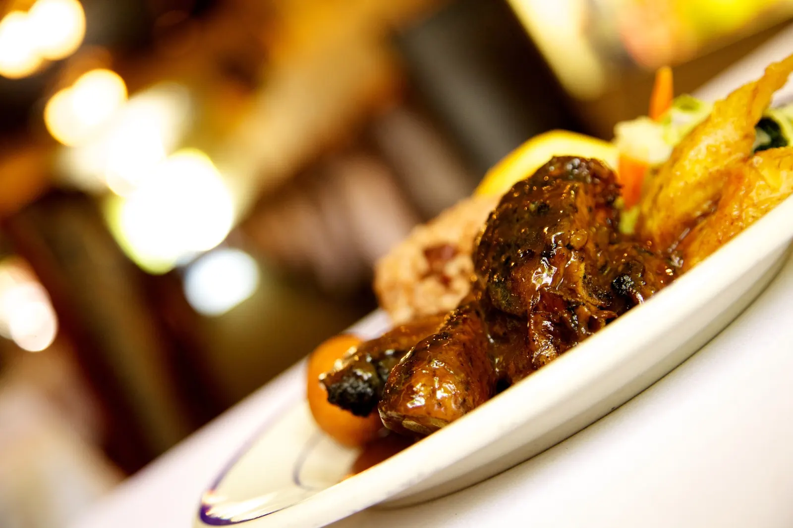 Glazed entree on a long white plate with warm restaurant lights behind it