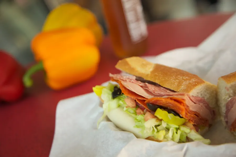 Italian sandwich in a paper-lined basket on a red table, with peppers behind it