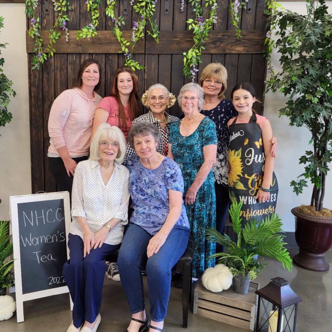 Women of New Hope Clovis dressed in semi-formal attire, posing for a photo at a Women's Tea event, with tables decorated with floral centerpieces in the background, at a key Christian Church in Clovis.
